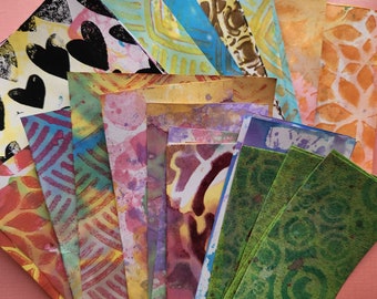 May include: A collection of rectangular paper bookmarks featuring various artistic techniques including black heart prints, geometric patterns, watercolor washes, and stenciled floral designs in a vibrant palette of blue, green, yellow, orange, and pink.