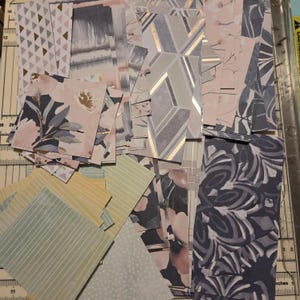 Pode incluir: Uma cole&ccedil;&atilde;o de retalhos de papel variados com diversos padr&otilde;es, incluindo formas geom&eacute;tricas, motivos florais, riscas e designs abstratos em tons de rosa, azul, cinzento, amarelo e branco, dispostos sobre uma base de corte com marcas m&eacute;tricas.