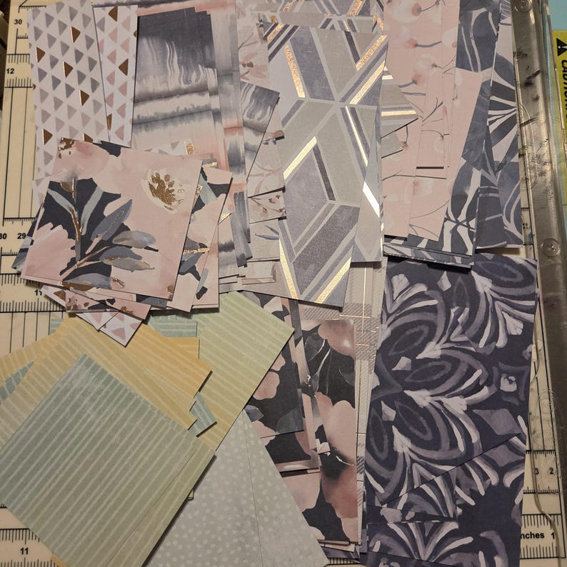 Pode incluir: Uma cole&ccedil;&atilde;o de retalhos de papel variados com diversos padr&otilde;es, incluindo formas geom&eacute;tricas, motivos florais, riscas e designs abstratos em tons de rosa, azul, cinzento, amarelo e branco, dispostos sobre uma base de corte com marcas m&eacute;tricas.