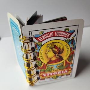 Può includere: Un piccolo taccuino realizzato con carte da gioco vintage rilegate con una spirale metallica dorata. La copertina presenta un ritratto circolare giallo di una persona con il testo Heraclio Fournier, Varios Premios e Vitoria.