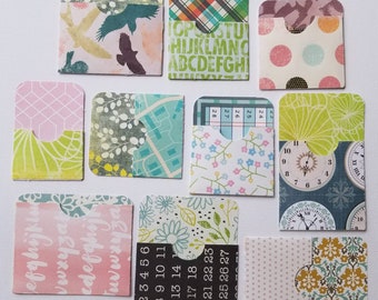 May include: Nine small, square, paper pockets with different patterns. The pockets have a cut-out shape at the top. The patterns include birds, alphabet letters, polka dots, a map, flowers, numbers, clocks, and a geometric design.
