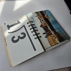 Può includere: Un calendario o libretto con rilegatura a spirale che presenta il numero 13 in un grande carattere nero su una pagina bianca, con il testo SEA WORLD visibile sul lato. La pagina adiacente mostra una fotografia a colori di un paesaggio urbano.