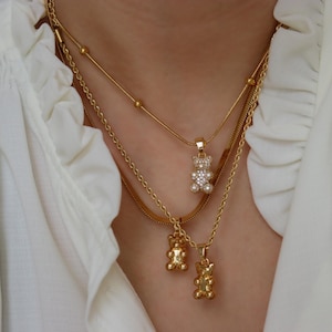 May include: Gold-toned necklaces layered on a white blouse. The necklaces feature delicate chains and bear-shaped pendants, one adorned with clear crystals. The jewelry adds a touch of sparkle and charm to the neckline.