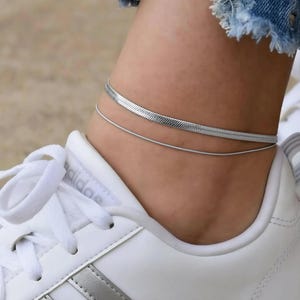 A close-up view of a person's ankle wearing a gold chain bracelet and white sneakers.