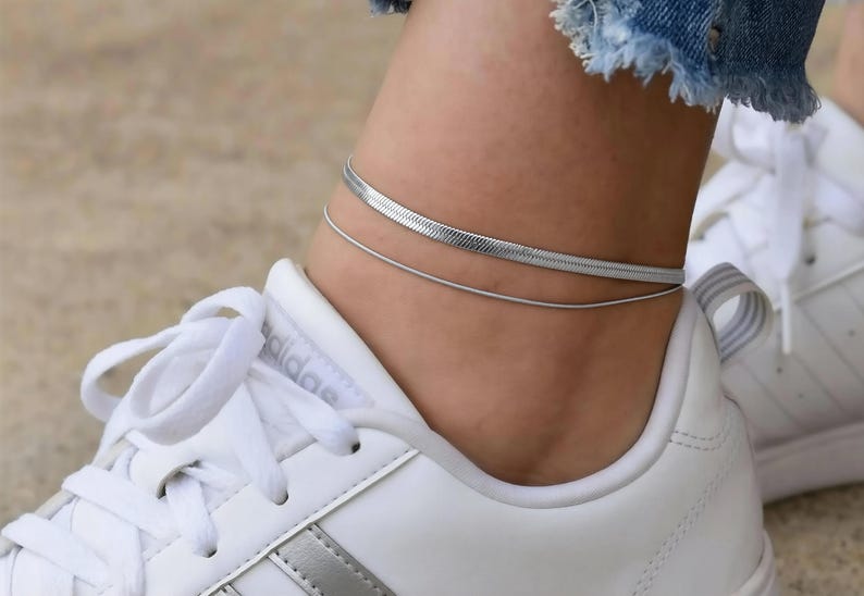 A close-up view of a person's ankle wearing a gold chain bracelet and white sneakers.