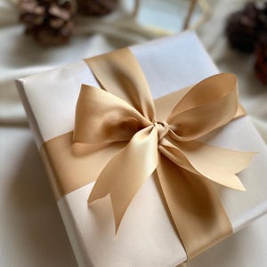 A gift box with a gold ribbon tied around it, placed on a white surface with pine cones in the background.