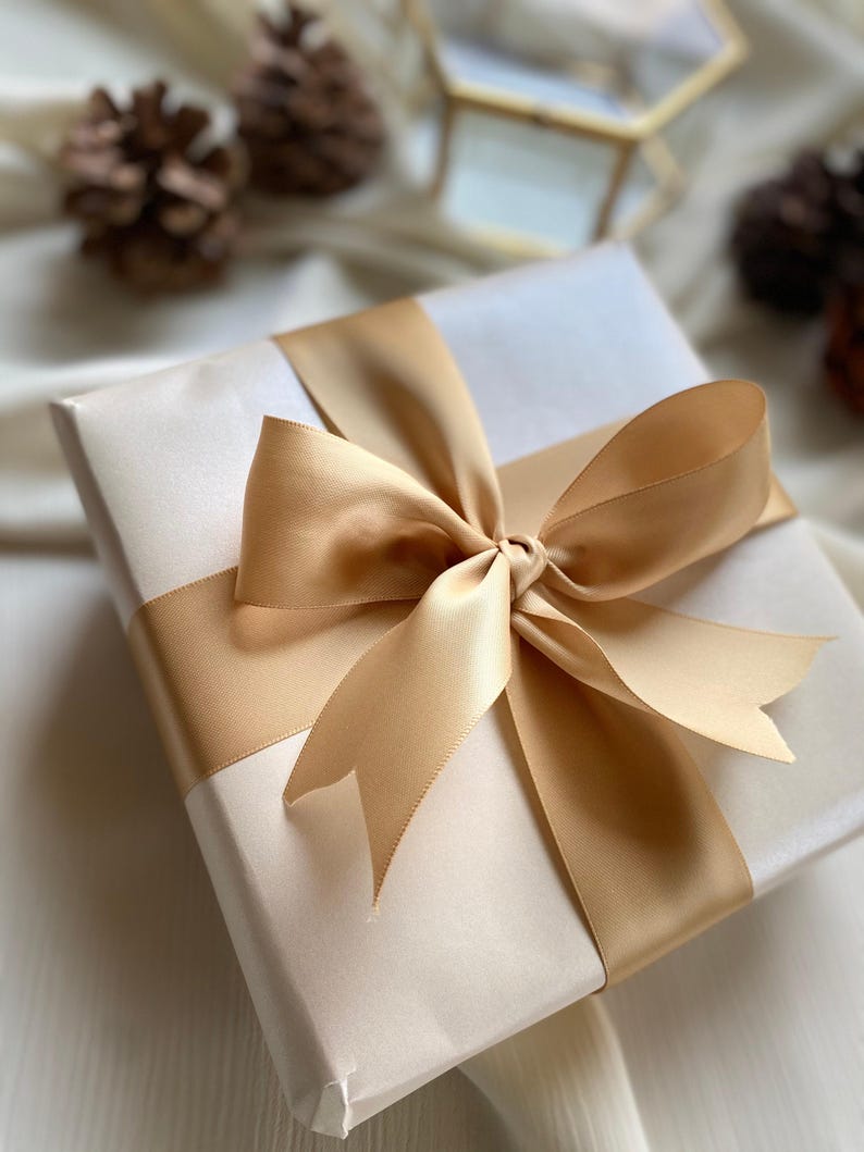 A gift box with a gold ribbon tied around it, placed on a white surface with pine cones in the background.