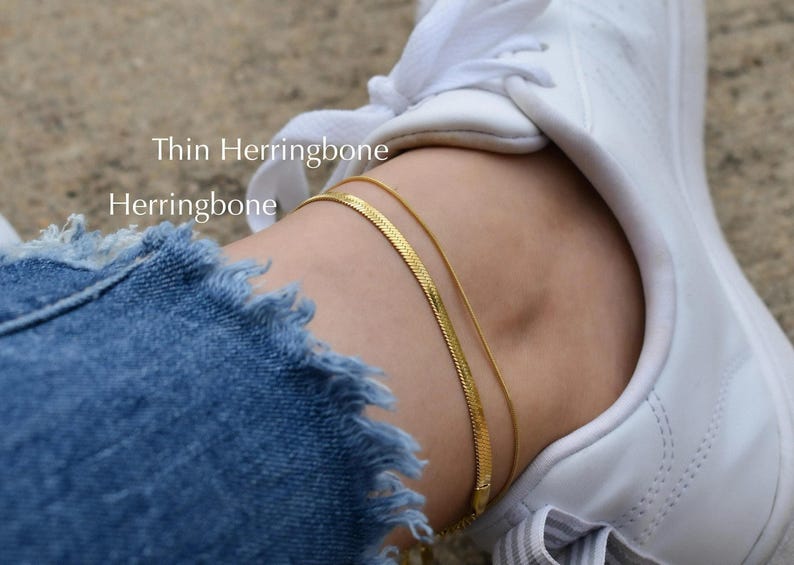 A close-up of a person's ankle wearing a gold chain bracelet and a white sneaker.