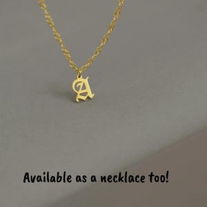 A gold-colored initial necklace pendant is displayed against a gray background.