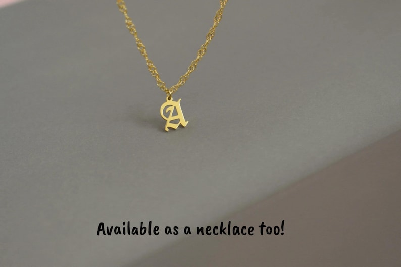 A gold-colored initial necklace pendant is displayed against a gray background.