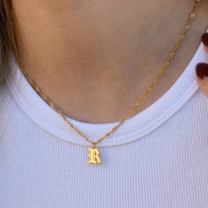 A close-up view of a person's neck and chest area, showing a gold necklace with a small pendant featuring the letter "R".