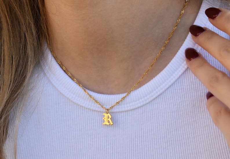 A close-up view of a person's neck and chest area, showing a gold necklace with a small pendant featuring the letter "R".