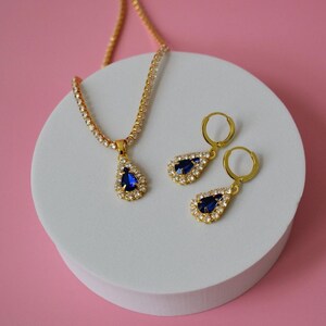 May include: A gold-toned jewelry set featuring a necklace and matching earrings. The necklace and earrings have a teardrop-shaped blue gemstone surrounded by small, clear crystals. The set is displayed on a white circular platform against a pink backdrop.
