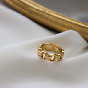 A gold ring with a chain-link design rests on a white fabric surface, with a gold-colored frame visible in the background.