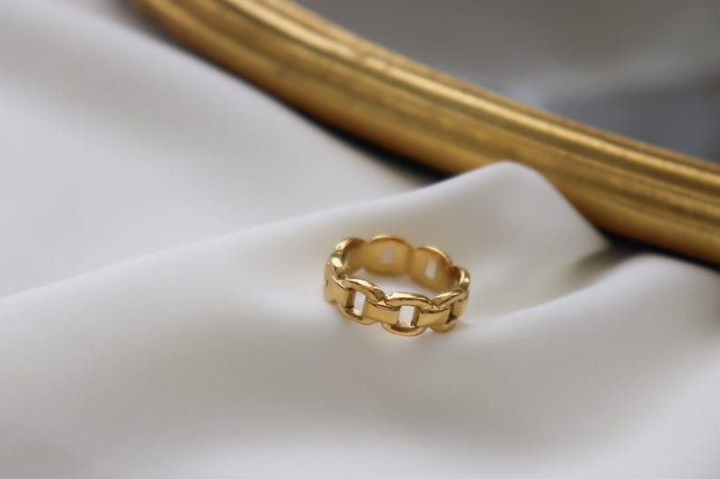 A gold ring with a chain-link design rests on a white fabric surface, with a gold-colored frame visible in the background.