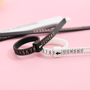 May include: A close-up shot of three ring sizers on a pink surface. Two are black with gold numbers, and one is white with black numbers. The ring sizers are used to measure ring sizes.