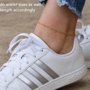 A close-up of a person's ankle wearing a gold ankle bracelet and white Adidas sneakers.