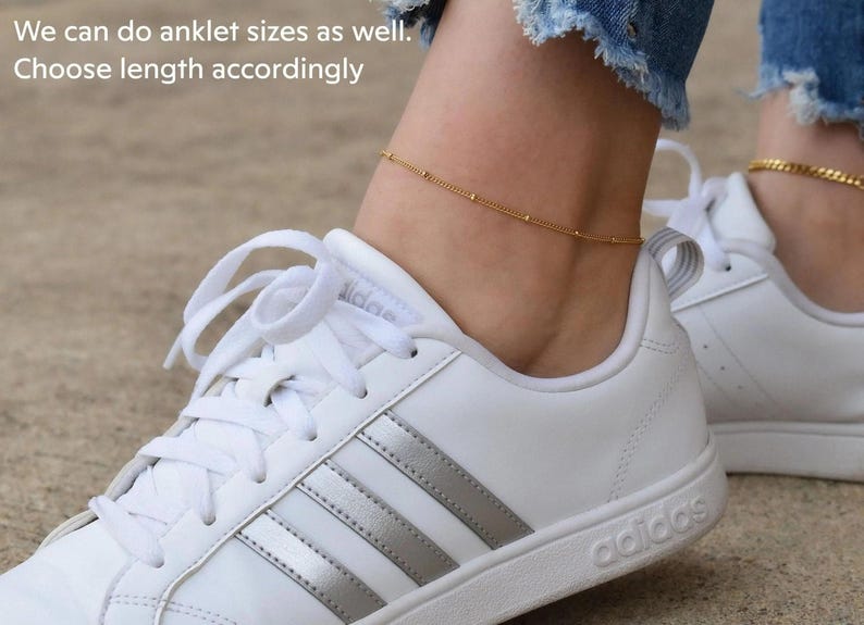 A close-up of a person's ankle wearing a gold ankle bracelet and white Adidas sneakers.