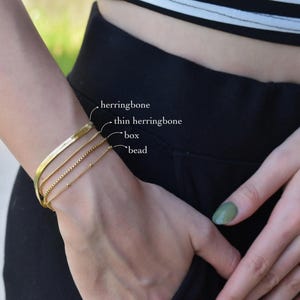 A close-up view of a person's wrist wearing a gold-colored bracelet with three distinct sections labeled as herringbone, thin herringbone, and bead.
