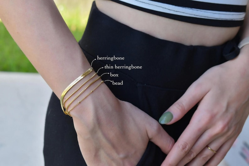A close-up view of a person's wrist wearing a gold-colored bracelet with three distinct sections labeled as herringbone, thin herringbone, and bead.