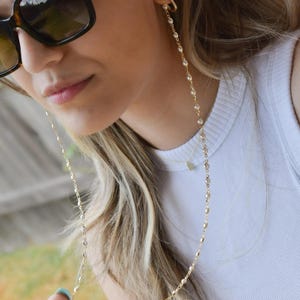 May include: Gold-toned eyeglass chain with small, clear beads. The chain is draped over the shoulder and attached to sunglasses. The text "EVIL EYE CHAIN" is visible at the bottom of the image.