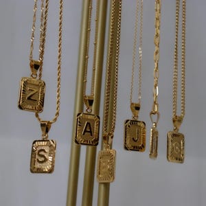 May include: Gold-colored initial necklaces with rectangular pendants. The pendants display the letters S, Z, A, J, E, and G. The necklaces hang from a gold display stand. The chains have different styles, including rope and box chains.