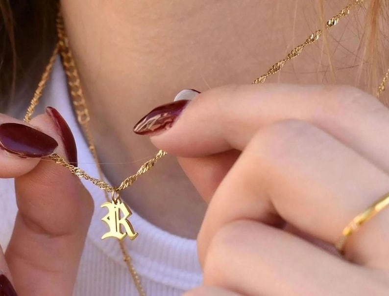 A close-up of a person's hands wearing a gold necklace and ring, with their nails painted in a burgundy color.