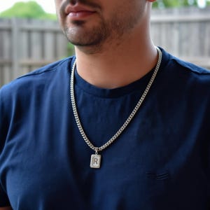 May include: A silver chain necklace with a square pendant featuring the letter "R" in a rectangular frame. The necklace is worn over a navy blue t-shirt.