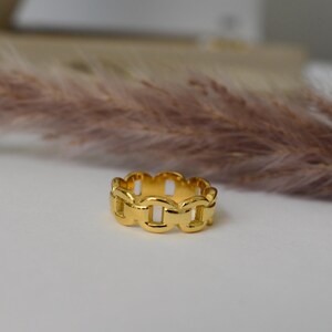 A gold ring with a unique chain design sits on a white surface, with a blurred background that includes a plant or flower.