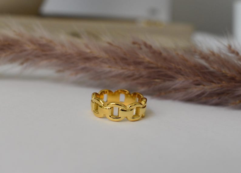 A gold ring with a unique chain design sits on a white surface, with a blurred background that includes a plant or flower.