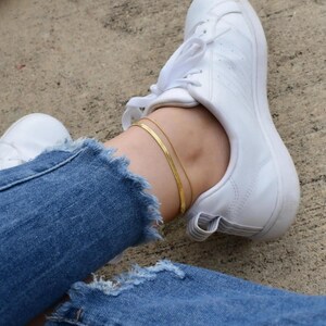 A person's legs wearing white sneakers and blue jeans, with a gold ankle bracelet visible.