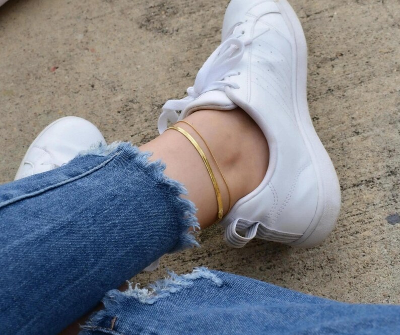 A person's legs wearing white sneakers and blue jeans, with a gold ankle bracelet visible.