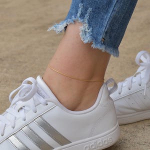 A close-up of a person's feet wearing white Adidas sneakers and gold ankle bracelets.