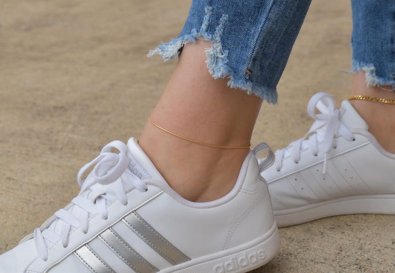 A close-up of a person's feet wearing white Adidas sneakers and gold ankle bracelets.