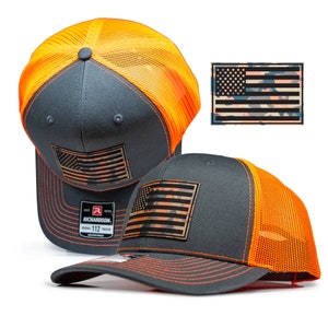 May include: Two gray and orange trucker hats with a camouflage American flag patch on the front. The hats are adjustable and have a mesh back.