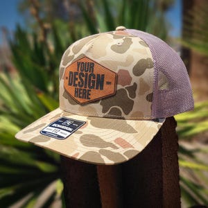 May include: A camouflage trucker hat featuring a tan and brown pattern with a brown leather-like hexagonal patch on the front. The patch displays the text YOUR DESIGN HERE in black. The hat has a mesh back and a sticker on the flat brim.