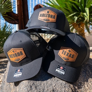 May include: Three black trucker hats with brown leather patches featuring the state outlines and names of Arizona, Nevada, and Texas. Each patch includes the state's founding year.