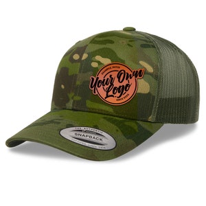 May include: A green camouflage trucker hat with a brown leather patch that says "Your Own Logo" in a circle. The hat has a mesh back and a snap closure.