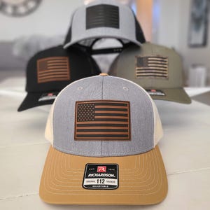 May include: A collection of Richardson 112 trucker hats with an American flag patch. The hats feature a variety of colors, including black, gray, and olive green, with brown and black flag designs. The front hat has a tan brim and a gray crown.