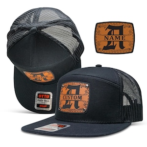 May include: Black trucker hat with a brown leather patch featuring a stylized letter "A" and the word "CUSTOM". The hat has a mesh back and a snap closure.