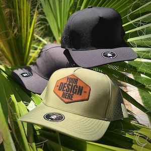 May include: Three baseball caps in black, gray, and olive green are displayed against a backdrop of green palm leaves. The olive green cap features a brown leather patch with the text "YOUR DESIGN HERE". Each cap has a curved brim and a small logo.