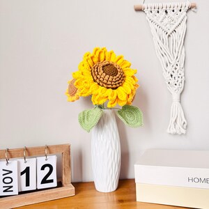 May include: A white ceramic vase with a textured surface holds a bouquet of crocheted sunflowers. The sunflowers are yellow with brown centers and green leaves. The vase is on a wooden table with a calendar that says "NOV 12" and a white box that says "HOME".