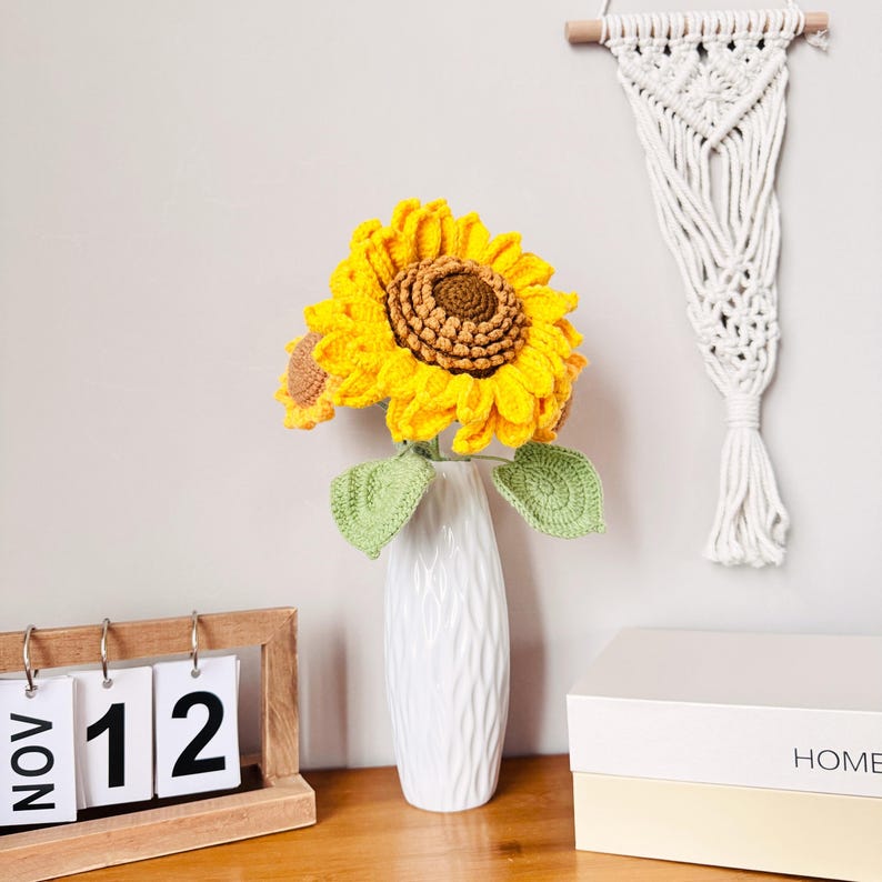 May include: A white ceramic vase with a textured surface holds a bouquet of crocheted sunflowers. The sunflowers are yellow with brown centers and green leaves. The vase is on a wooden table with a calendar that says "NOV 12" and a white box that says "HOME".