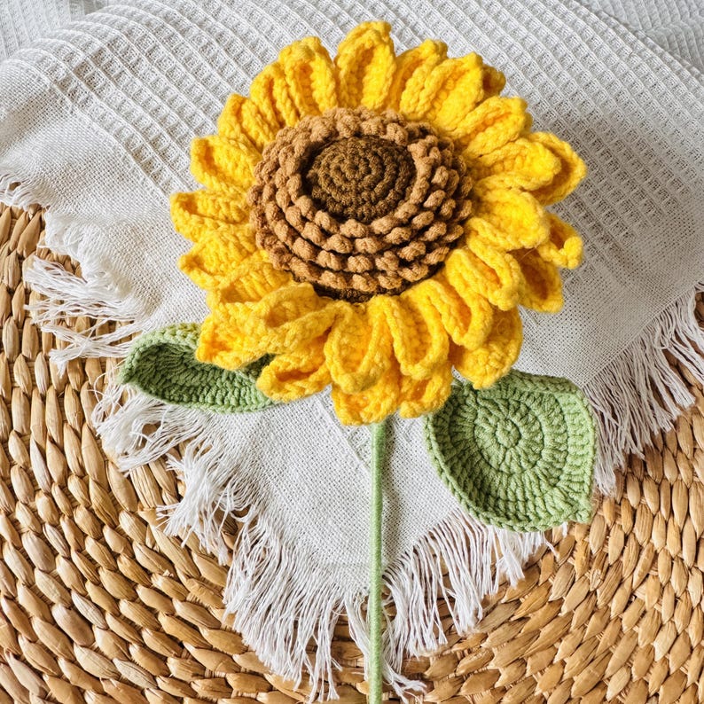 May include: A crocheted sunflower with yellow petals, a brown center, and green leaves. The sunflower is sitting on a white crocheted fabric with fringe.