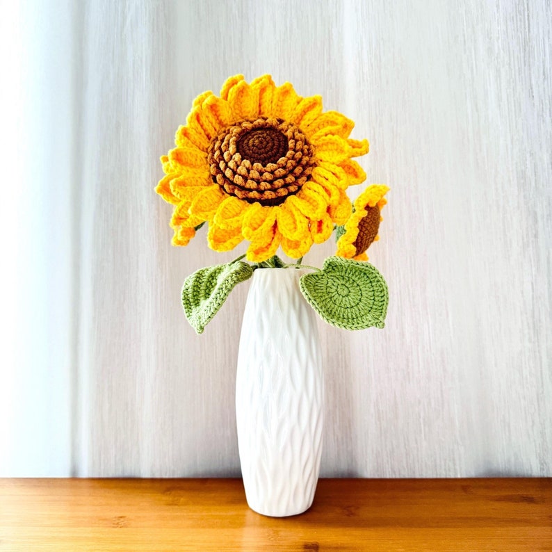 May include: A crocheted sunflower bouquet in a white ceramic vase. The sunflower is yellow with a brown center and green leaves.