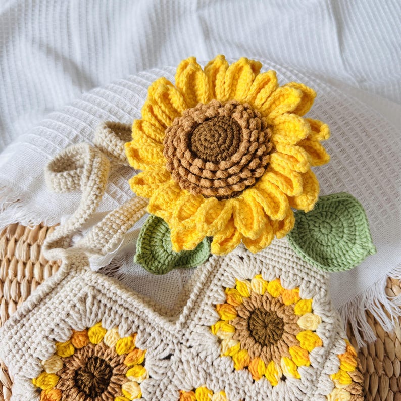 May include: A crocheted sunflower bag with a brown and yellow sunflower design. The bag has a long strap and is made of beige yarn.