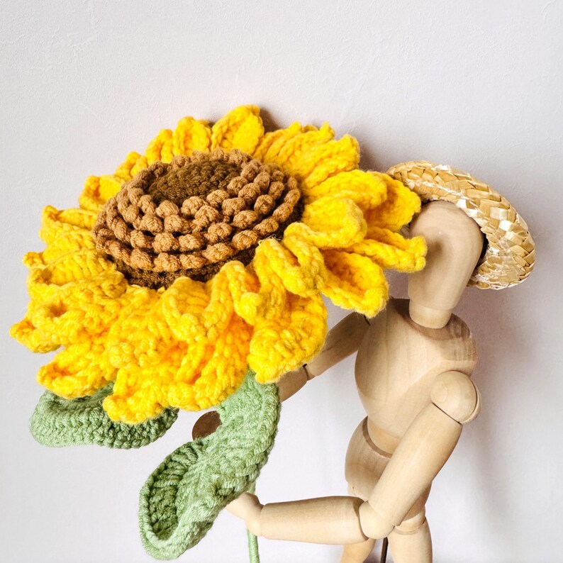 May include: A crocheted sunflower with a brown center and yellow petals. The sunflower is held by a wooden mannequin wearing a straw hat.