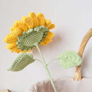 May include: A crocheted sunflower with yellow petals and green leaves. The sunflower is standing upright in a wicker basket.
