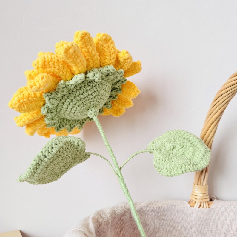 May include: A crocheted sunflower with yellow petals and green leaves. The sunflower is standing upright in a wicker basket.