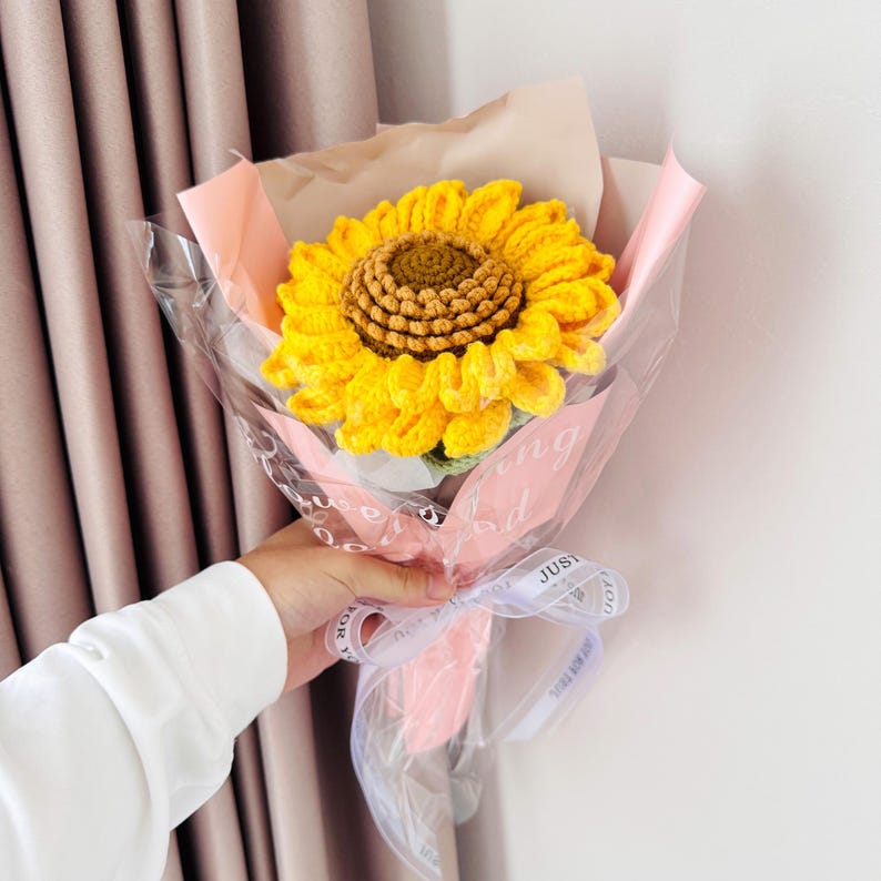 May include: A crocheted sunflower bouquet in a pink and white wrapping with a clear plastic sleeve. The bouquet is tied with a white ribbon with the text "Just for You" printed on it.